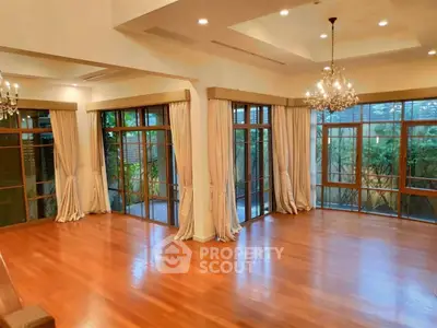 Spacious living room with hardwood floors and elegant chandeliers, featuring large windows and lush garden view.