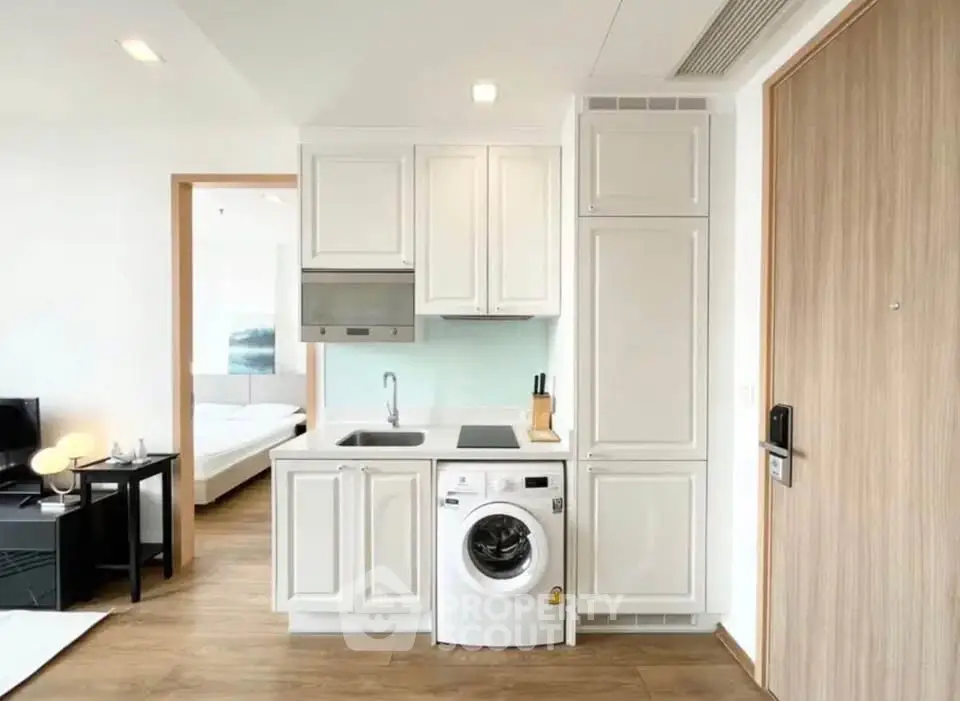 Modern studio apartment with compact kitchen and washing machine, featuring sleek cabinetry and wood flooring.