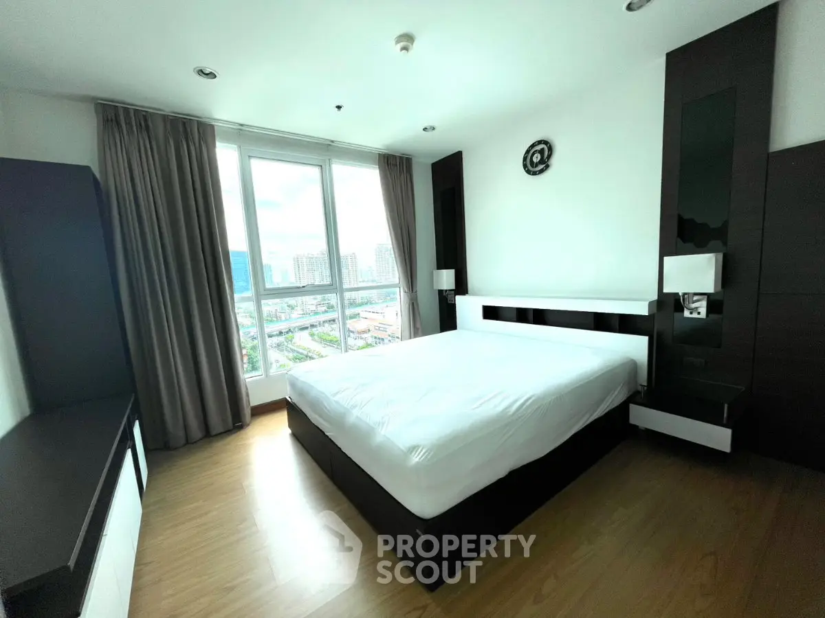 Modern bedroom with large window offering city view, featuring sleek furniture and wooden flooring.