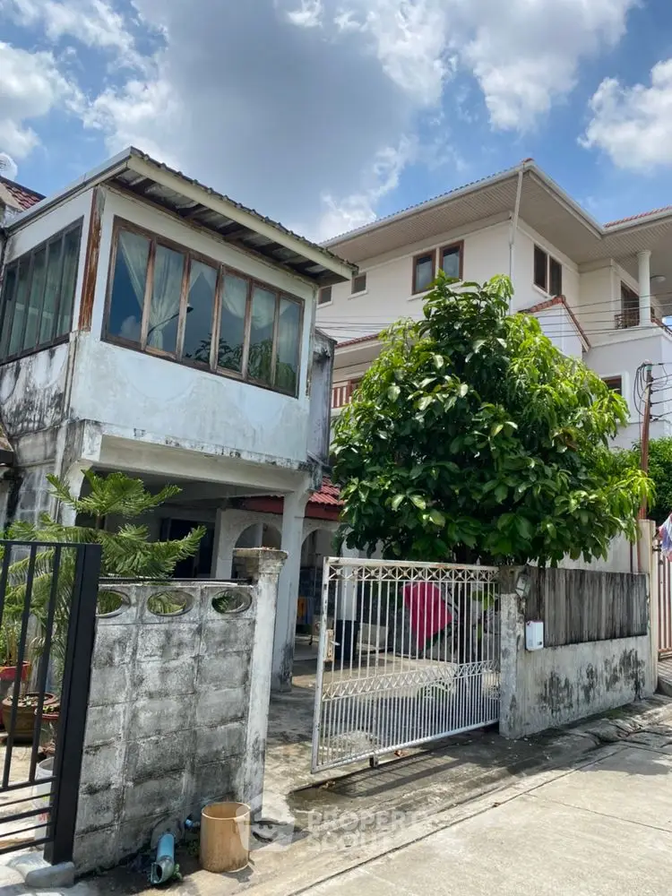 Charming two-story house with gated entrance and lush greenery under a bright blue sky.