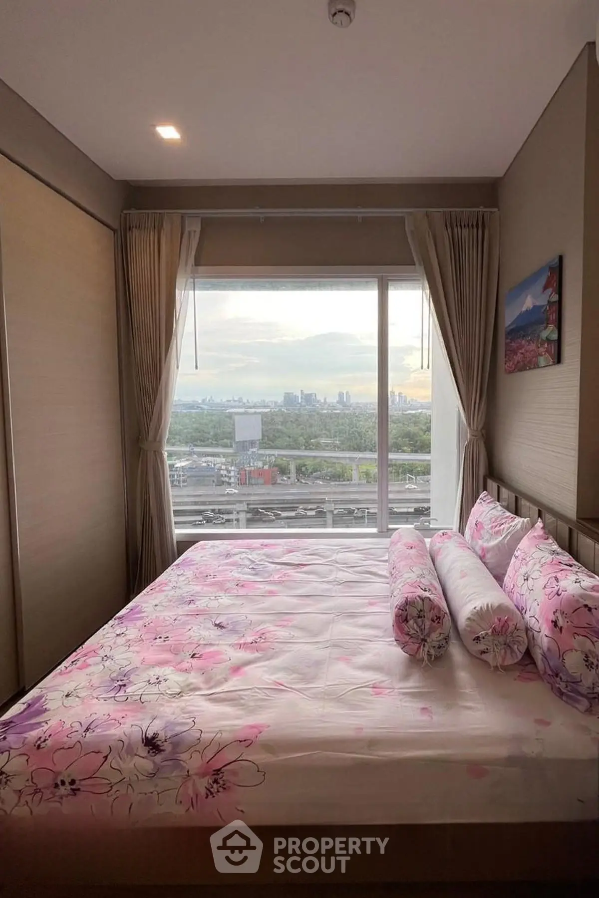 Cozy bedroom with large window offering stunning cityscape views, featuring floral bedding and elegant curtains.