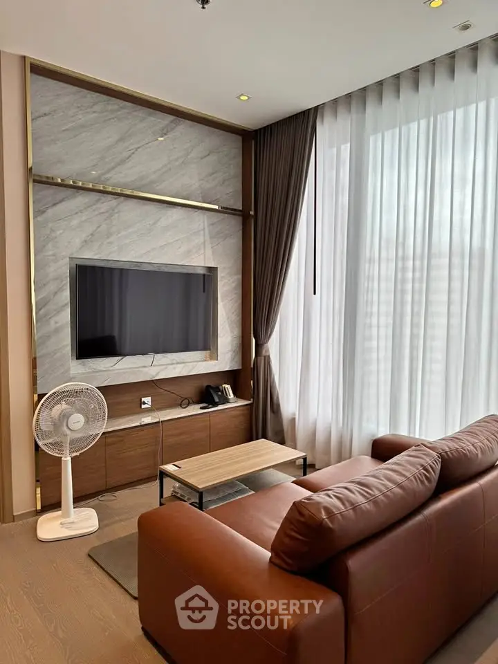 Modern living room with leather sofa, wall-mounted TV, and large window with sheer curtains.