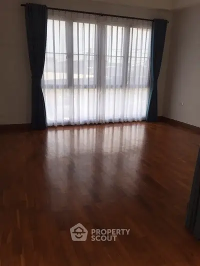 Spacious empty room with wooden flooring and large windows with curtains.