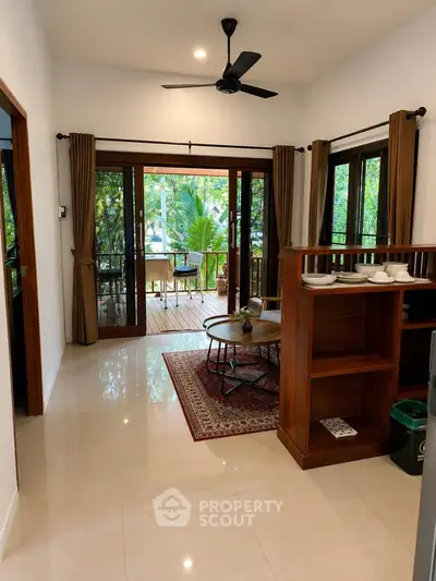 Charming living room with large windows opening to a lush balcony, featuring elegant wooden furniture and ceiling fan.
