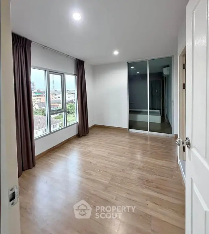 Spacious empty bedroom with wooden flooring and large windows offering city views.