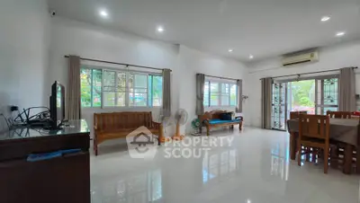 Spacious living room with large windows and natural light, featuring wooden furniture and tiled flooring.