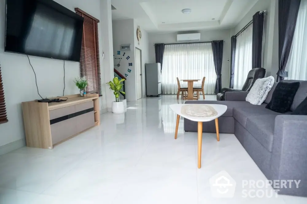 Spacious and modern living room with glossy white floors, elegant gray sofa set, and a large flat-screen TV, leading to a cozy dining area with natural light.