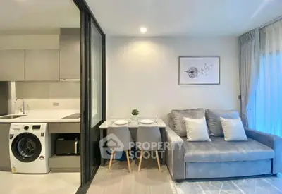 Modern living room with compact kitchen and washing machine, featuring cozy gray sofa and dining area.