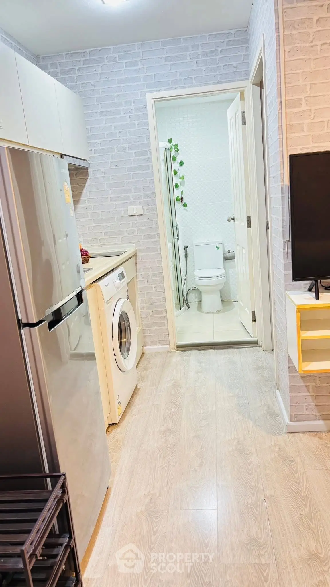 Modern kitchen with washing machine and fridge leading to a bathroom, featuring sleek design and light wood flooring.