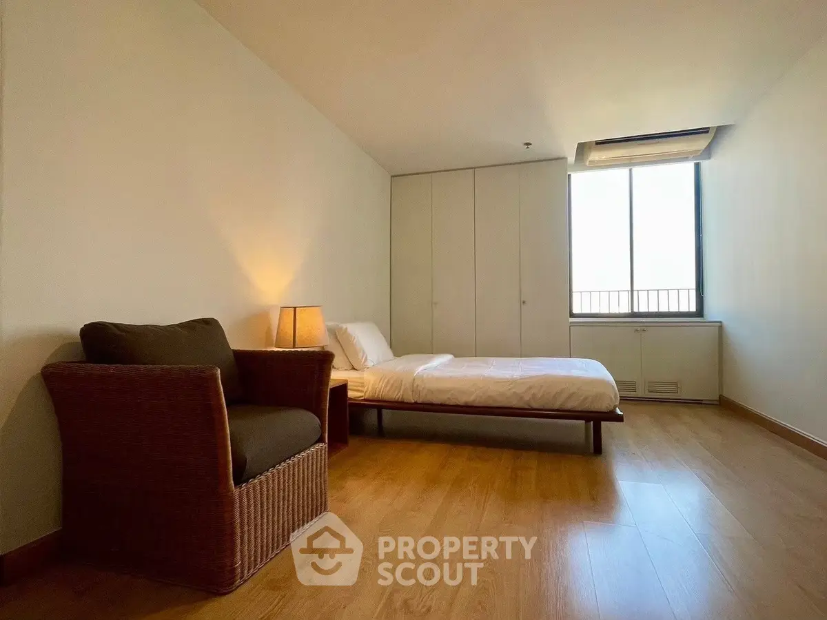 Spacious bedroom with wooden flooring and cozy wicker chair, featuring a large window for natural light.