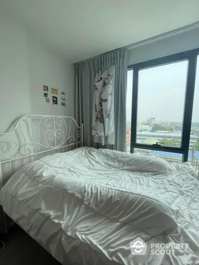 Cozy bedroom with large window offering city view, featuring white bed and decorative curtains.