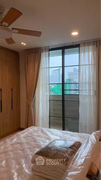 Cozy bedroom with city view and large window, featuring modern wooden wardrobe and ceiling fan.