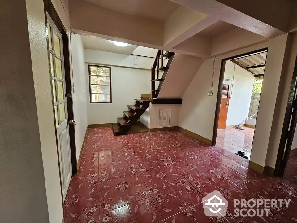 Spacious living area with striking red tiled flooring and an elegant wooden staircase leading to the upper level, surrounded by natural light from multiple windows.