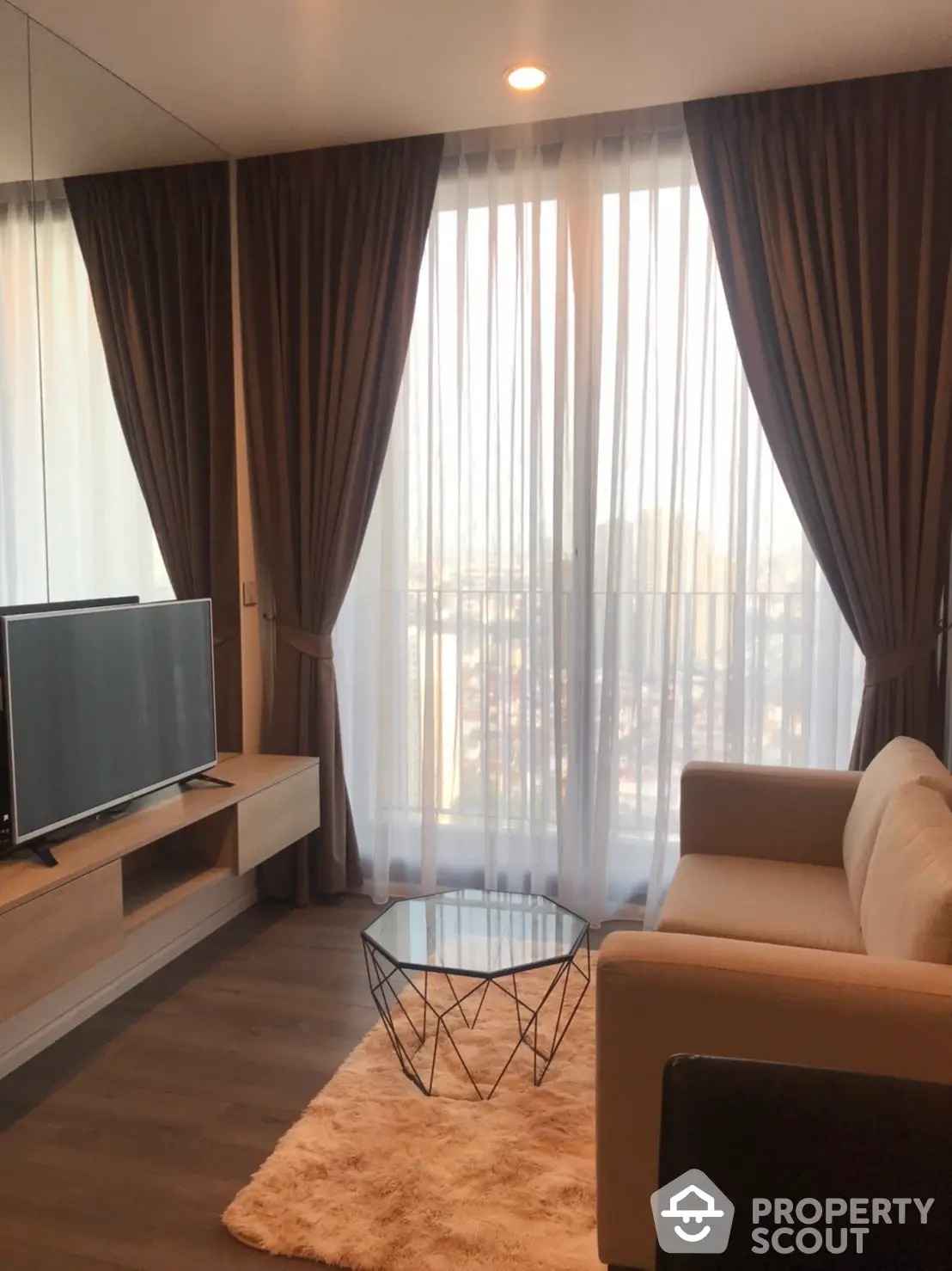 Cozy living room with warm wooden floors, plush sofa, and floor-to-ceiling curtains framing a sunlit city view, perfect for urban living.