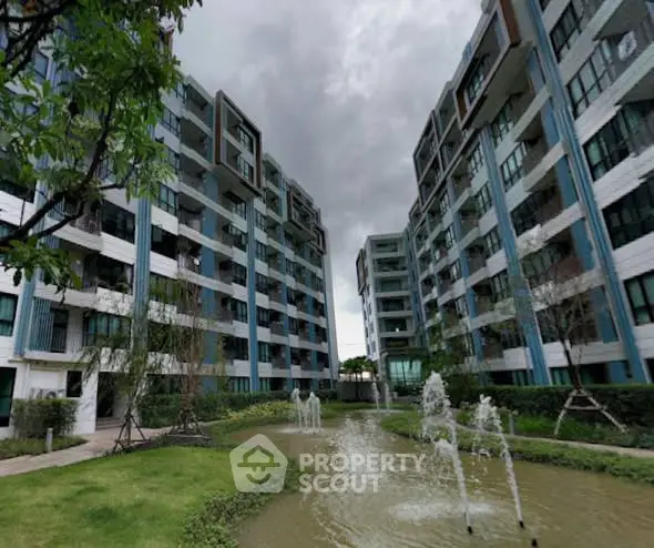 Modern apartment complex with lush garden and water feature under cloudy sky.