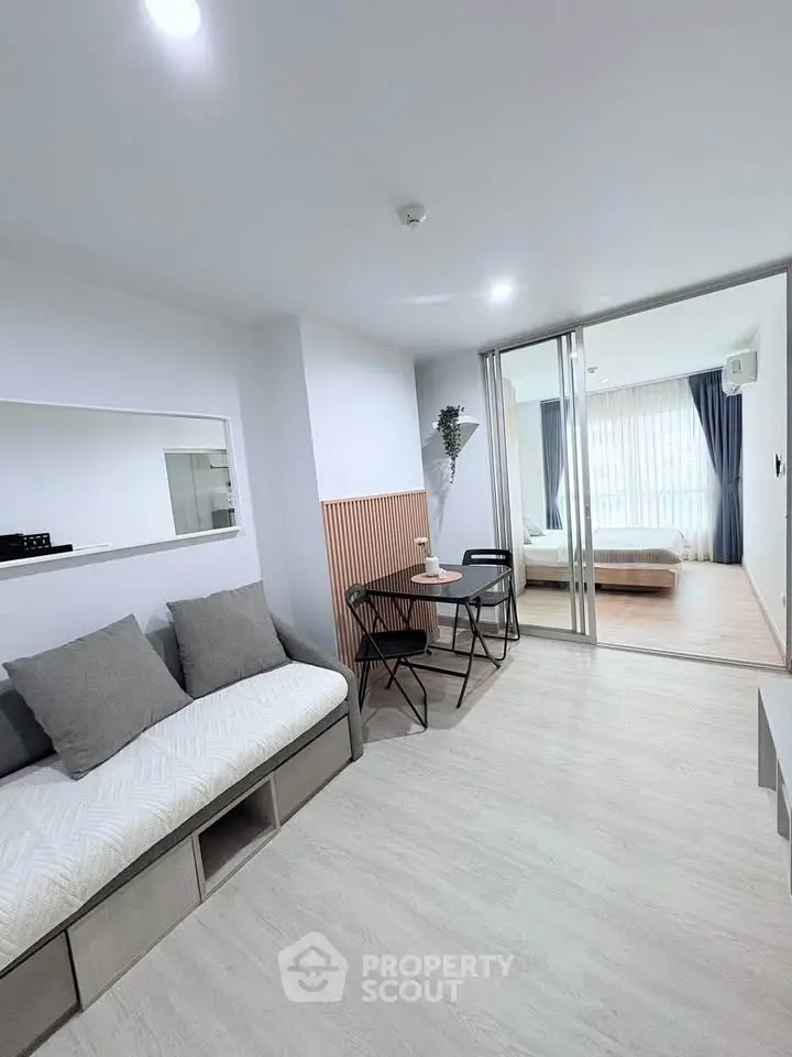 Modern open-plan living room with cozy seating and dining area, leading to a bright bedroom with large windows.