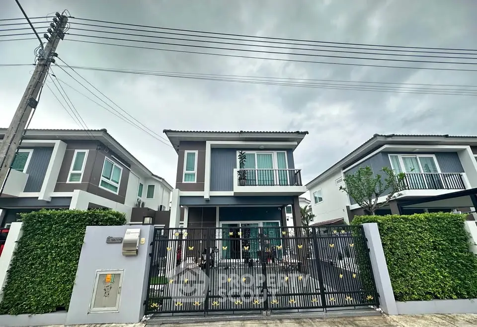 Modern two-story house with gated entrance and lush greenery under cloudy skies.