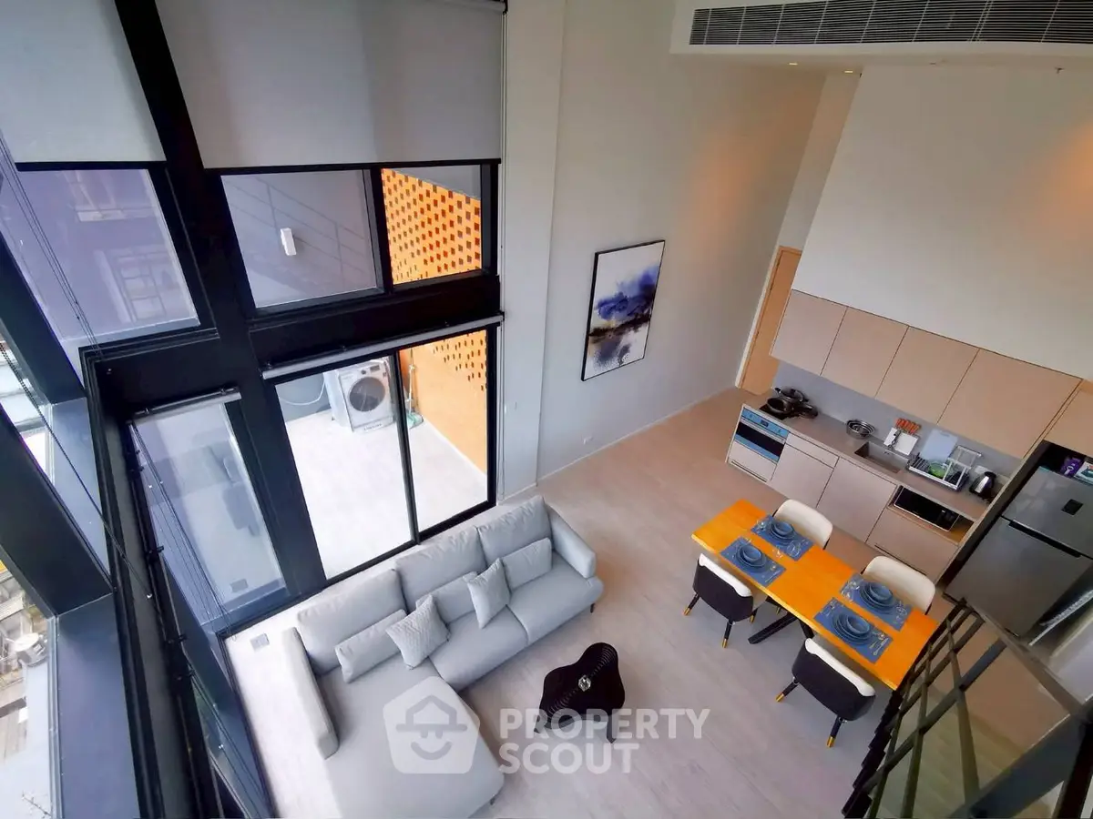 Modern loft-style living room with high ceilings and open kitchen layout, featuring sleek furniture and large windows.