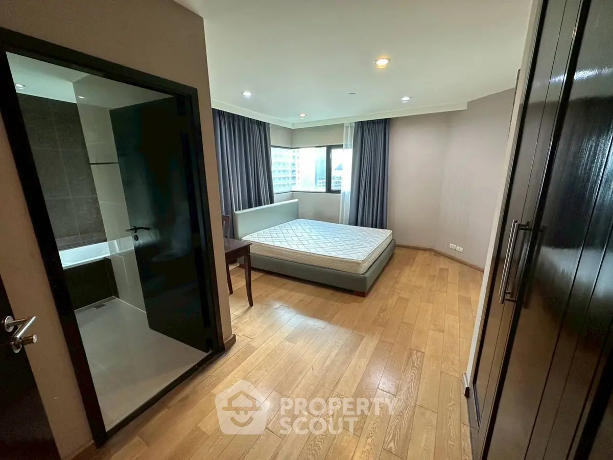 Spacious bedroom with wooden flooring and large window offering city views, adjacent to modern bathroom.