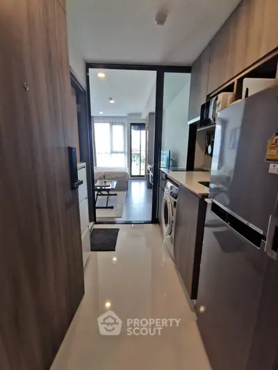 Modern apartment hallway with sleek kitchen and washing machine, leading to a bright living area with large windows.