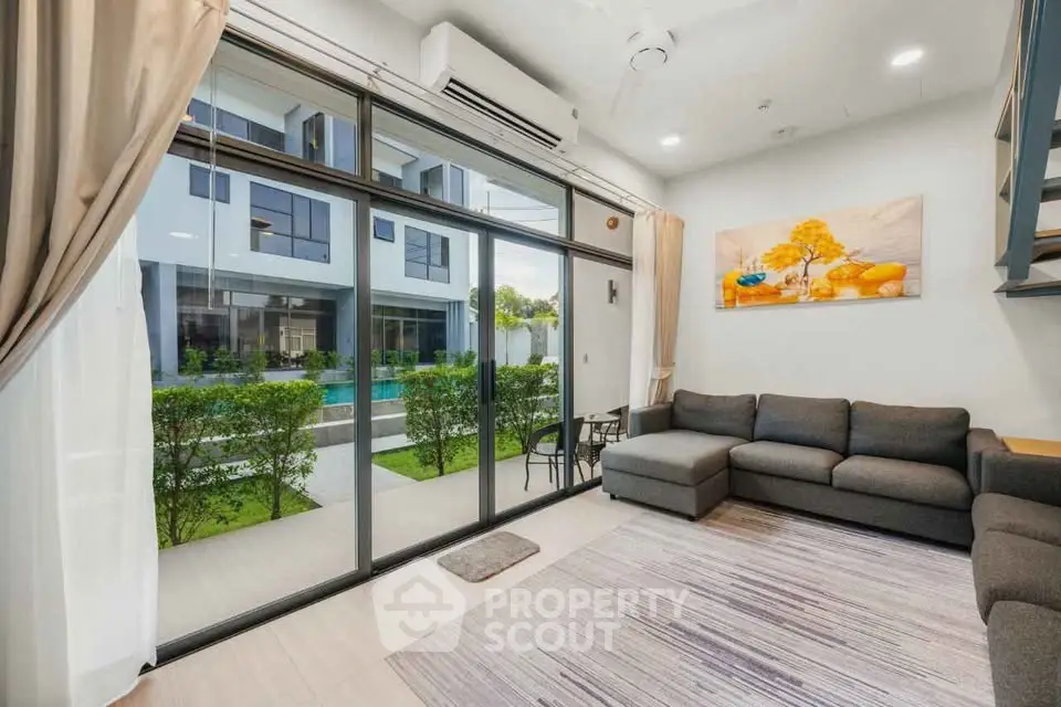 Modern living room with large windows overlooking garden and pool, featuring stylish gray sectional sofa.