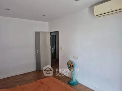 Minimalist bedroom with air conditioning and wooden flooring, featuring a simple fan and orange bedding.