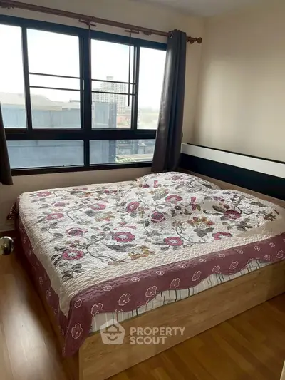 Cozy bedroom with large window offering city views, featuring a floral bedspread and wooden flooring.