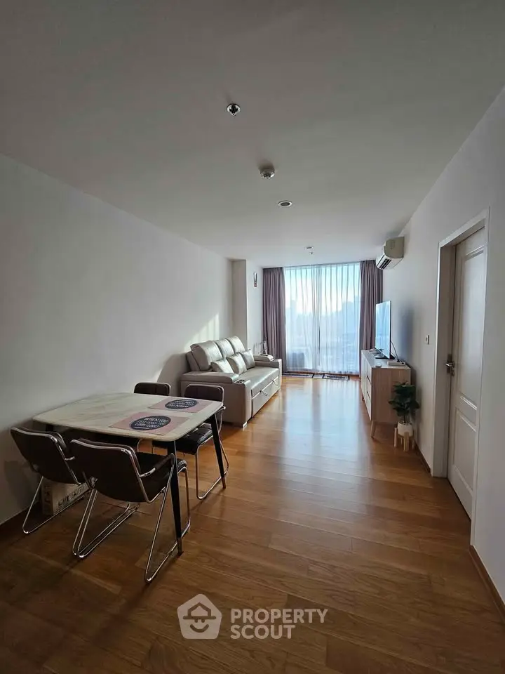 Spacious living room with wooden flooring and modern furniture, featuring a large window with natural light.