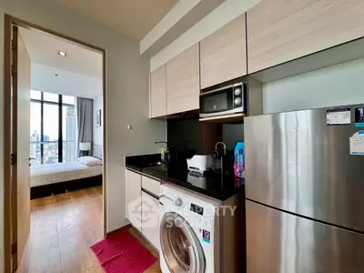 Modern apartment kitchen with washing machine and view into bedroom, featuring sleek cabinetry and stainless steel appliances.