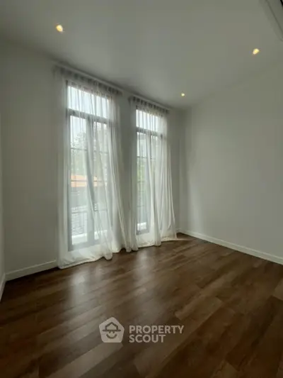 Spacious and well-lit living room with elegant hardwood floors and large windows draped with sheer curtains, offering a serene ambiance.