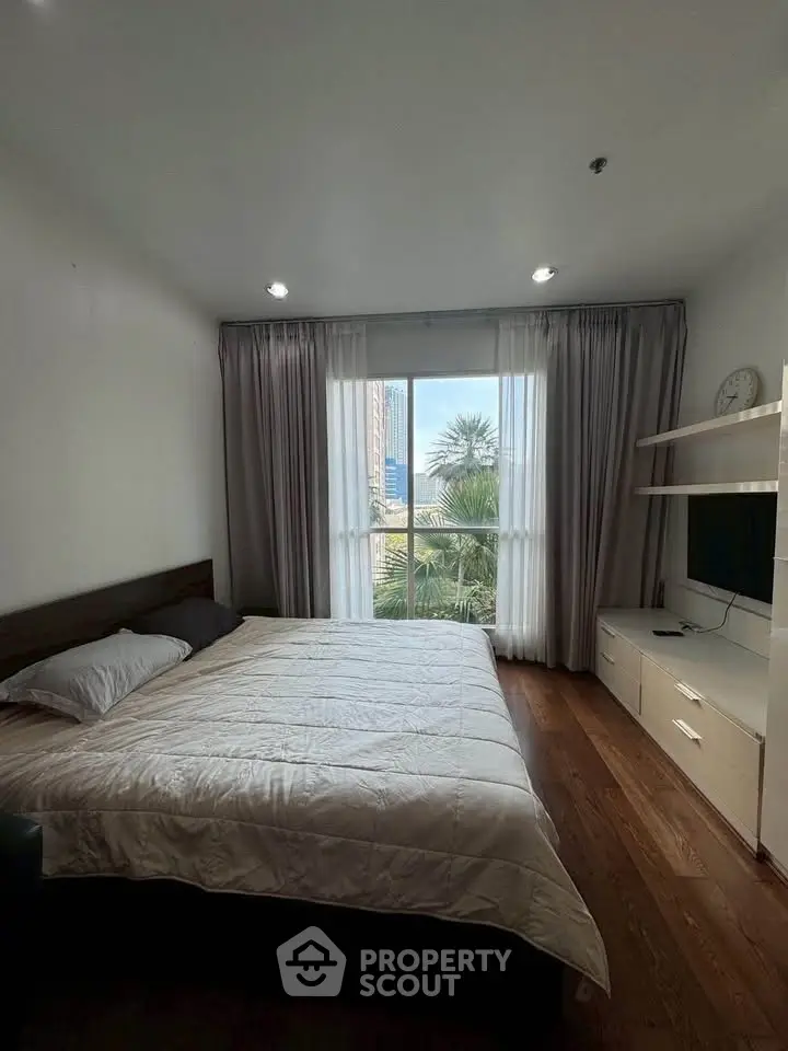 Spacious bedroom with large window offering city view, featuring modern furnishings and wooden flooring.