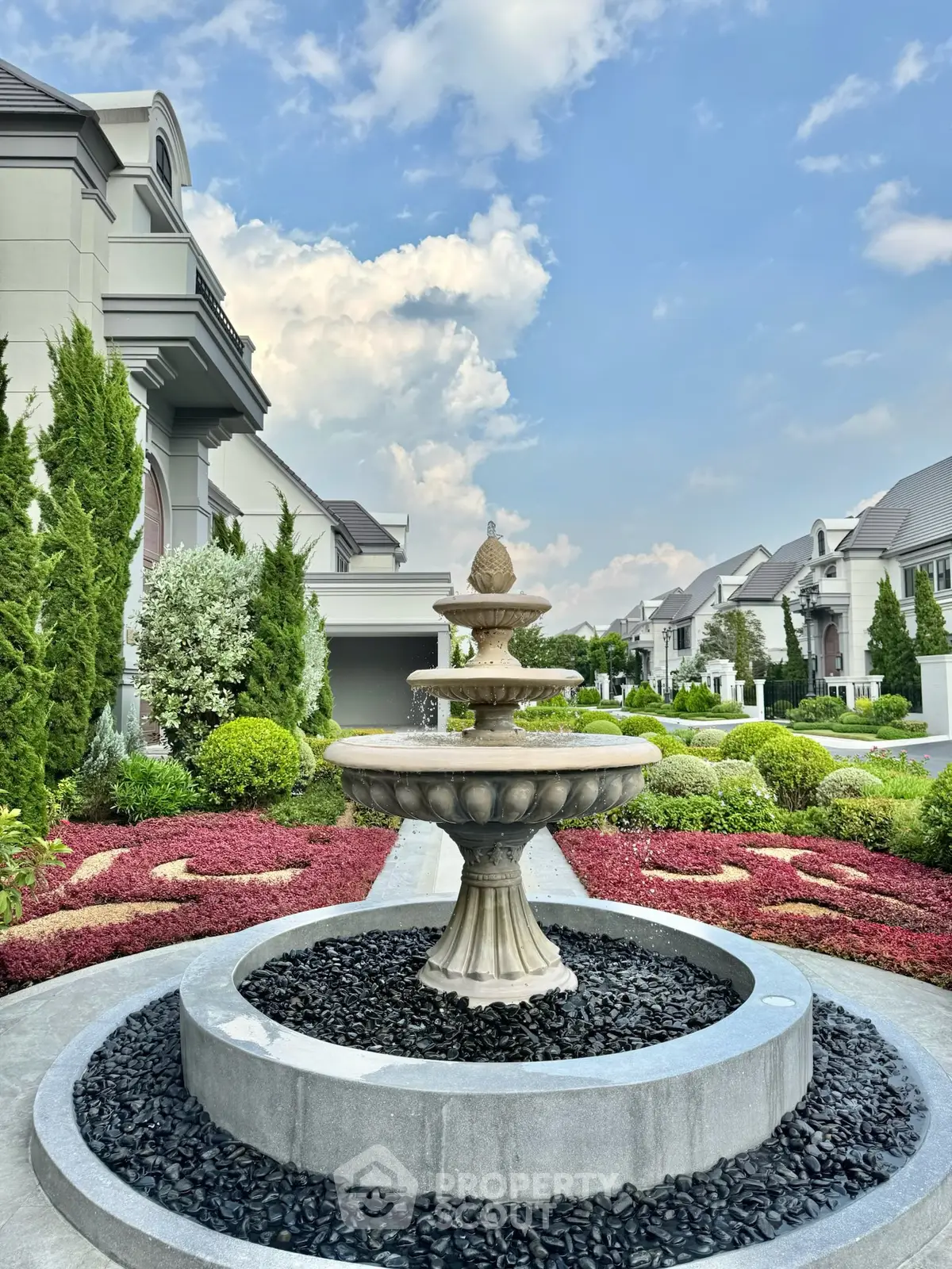 Luxurious residential entrance with elegant fountain and manicured gardens under a clear blue sky.