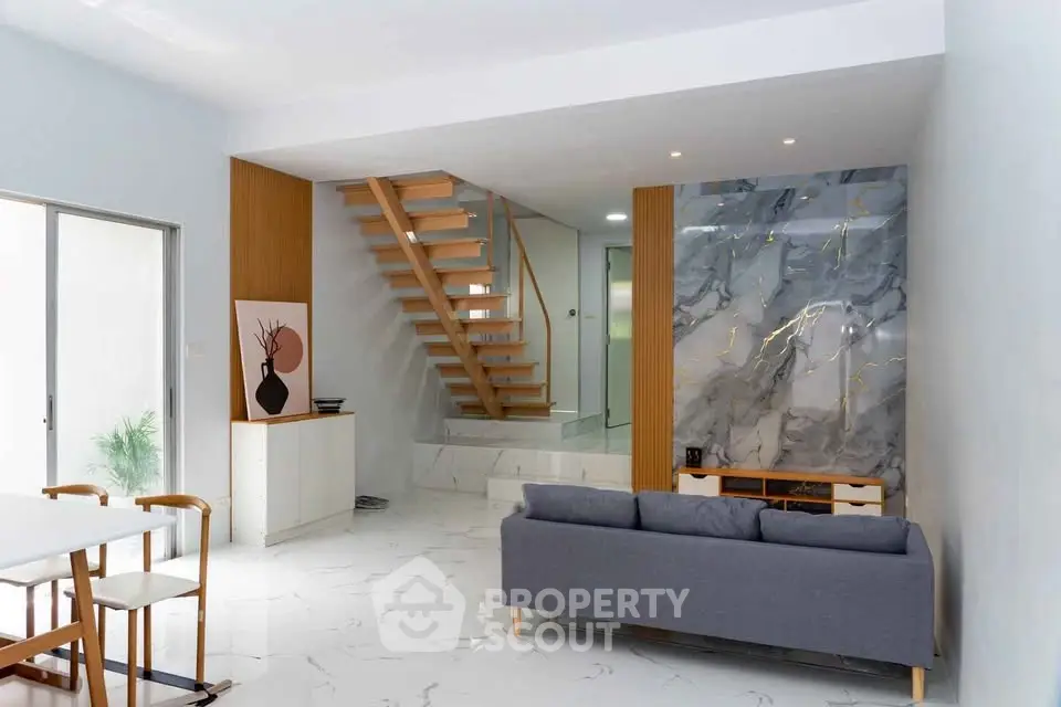 Modern living room with stylish staircase and marble accent wall, featuring sleek furniture and natural light.