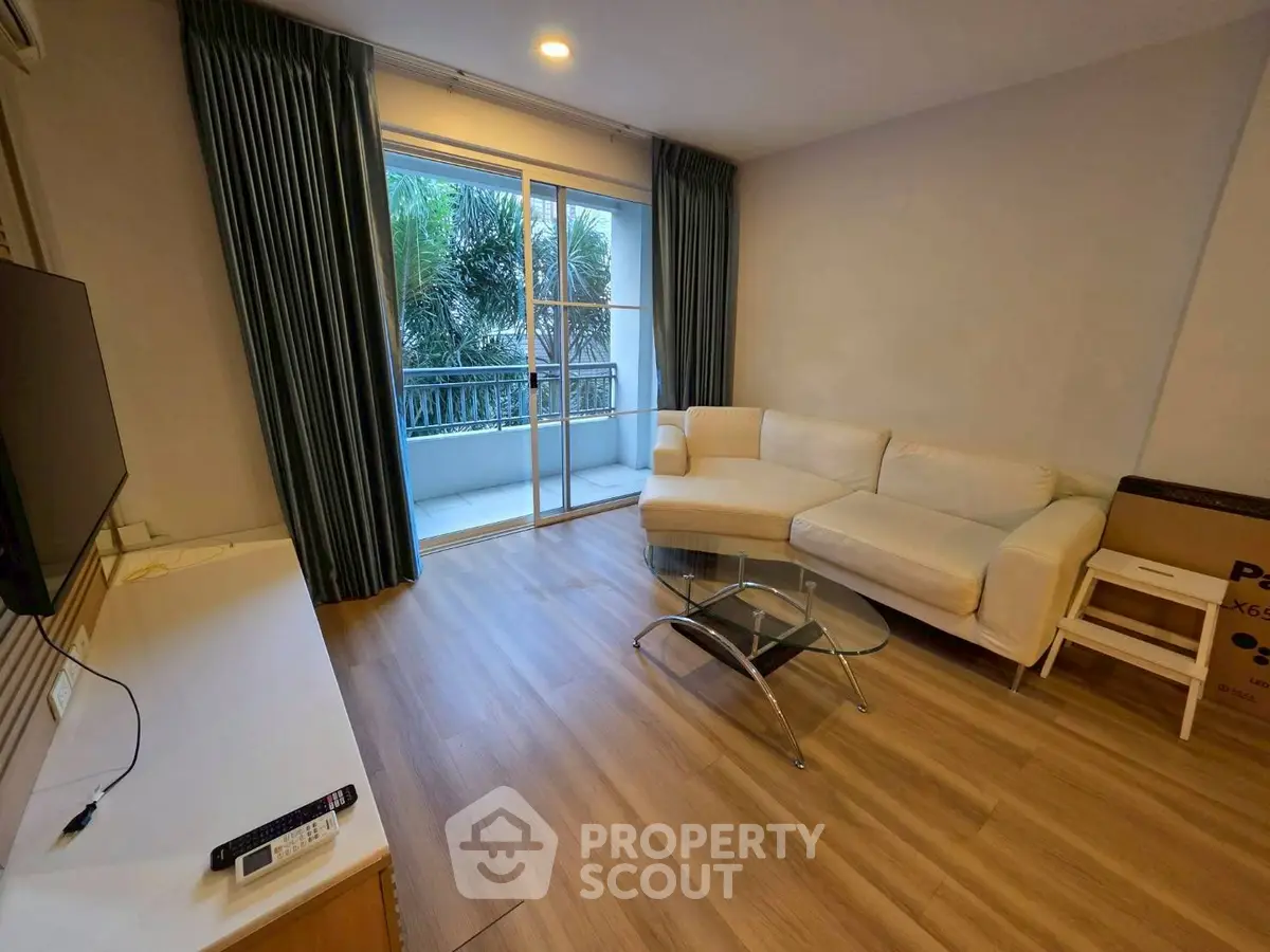 Modern living room with white sofa and glass coffee table, opening to a balcony with lush greenery view.