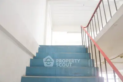 Brightly lit stairway with blue steps and red handrails leading to an airy upper level, offering a glimpse into the building's interior design.