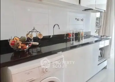 Modern kitchen with sleek black countertop and washing machine, featuring a fruit basket and large window.