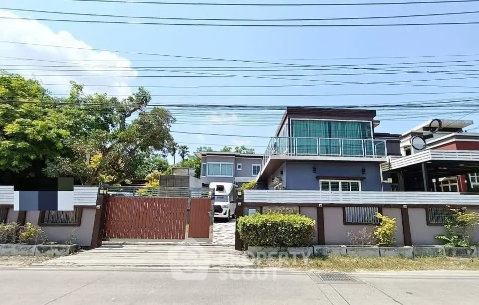 Modern two-story house with large windows and gated entrance, surrounded by lush greenery.
