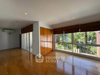 Spacious living room with hardwood floors and large windows overlooking lush greenery.