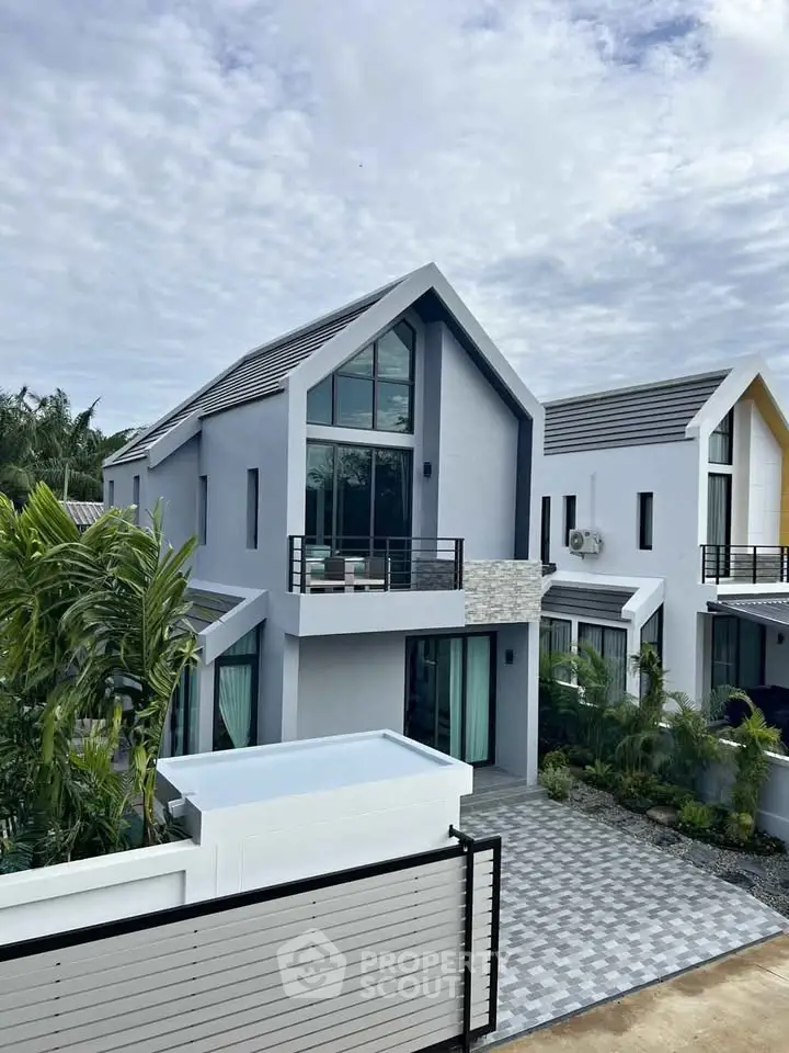 Modern two-story house with sleek design and large windows, surrounded by lush greenery and a paved driveway.