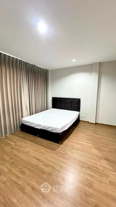 Spacious bedroom with wooden flooring and large window draped with curtains, featuring a comfortable bed.