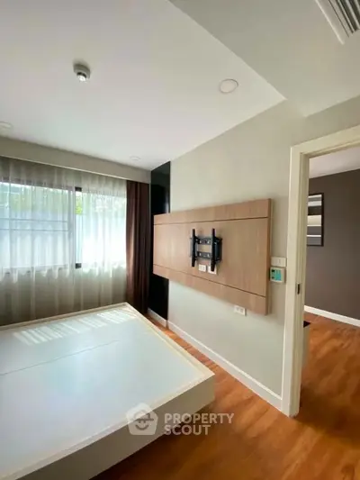 Modern bedroom with wooden flooring and large window, featuring a wall-mounted TV setup.