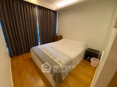 Cozy bedroom with wooden flooring and large window draped with curtains, featuring a comfortable bed and nightstand.