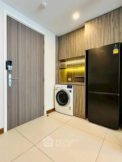 Modern apartment entryway with sleek wooden door, washing machine, and refrigerator in compact kitchen area.