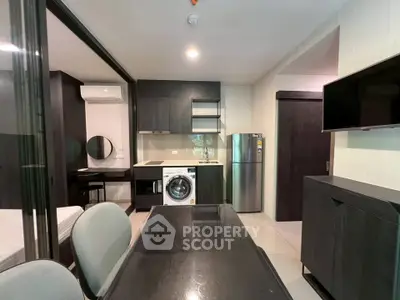 Modern apartment interior with kitchen, dining area, and washing machine, featuring sleek dark cabinetry.