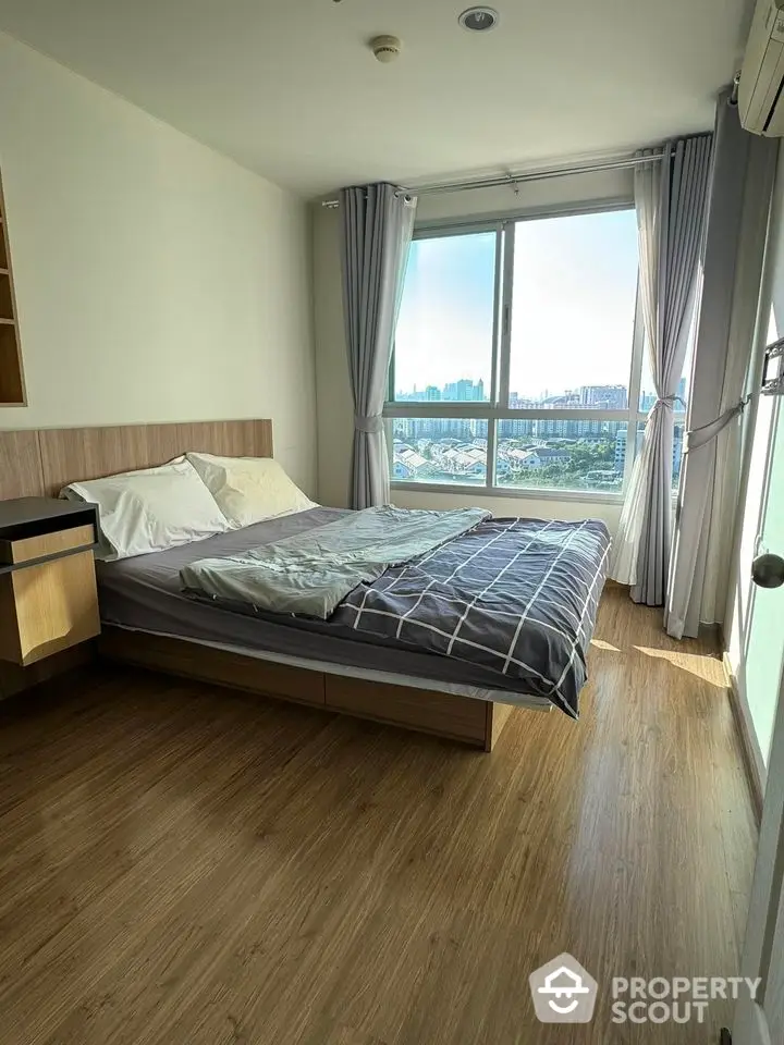 Bright bedroom with large window and city view, featuring modern wooden flooring and cozy bedding.