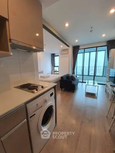 Modern open-plan living room with kitchen and washing machine, featuring large windows and balcony access.