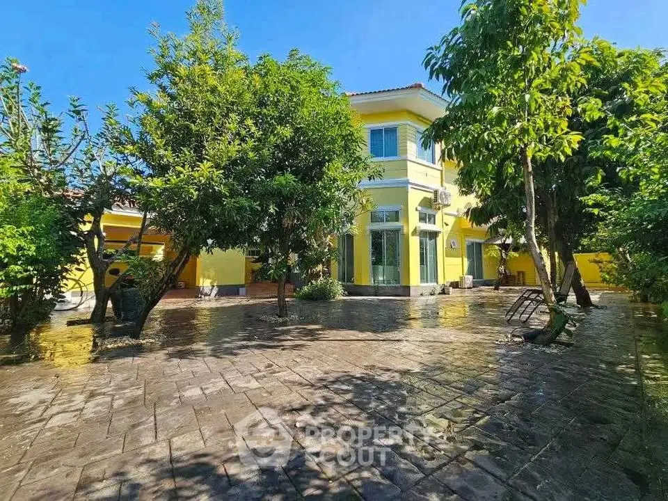 Charming yellow house with lush garden and spacious paved courtyard under clear blue sky.