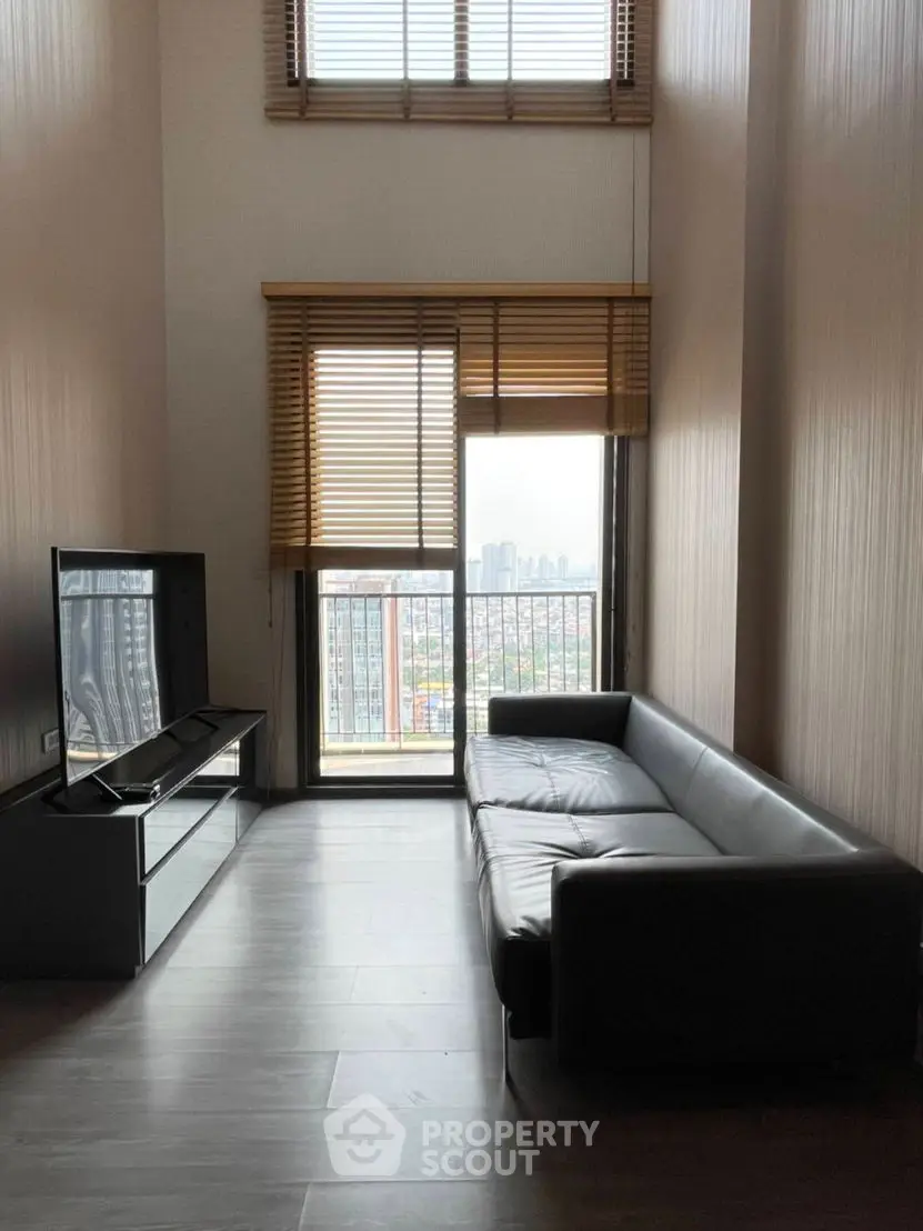 Modern living room with high ceiling and city view, featuring sleek black sofa and large windows.