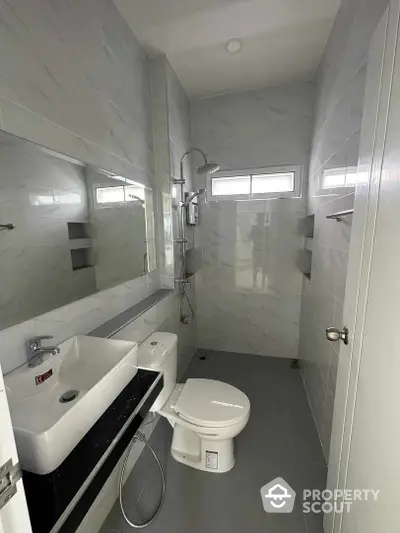 Sleek modern bathroom with glossy marble tiles, wall-mounted sink, and a spacious shower area, illuminated by natural light from frosted windows.