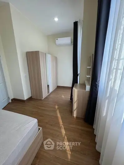 Bright bedroom with wooden flooring and modern furniture, featuring a large window with curtains.